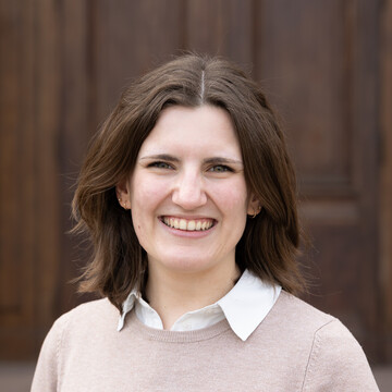 Katharina Schmidt. Sie trägt einen beigen Pullover über einem weißen Hemd und steht vor einer Holztür.