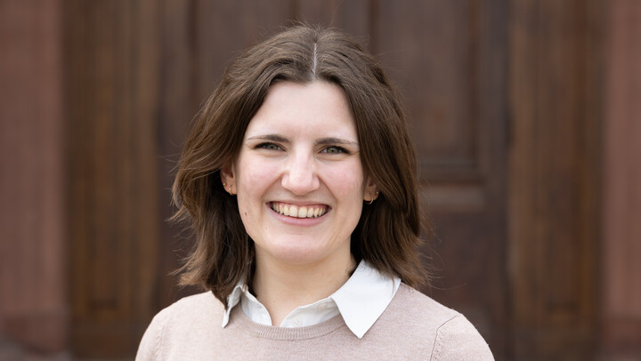 Katharina Schmidt. She is wearing a beige sweater over a white shirt and is standing in front of a brown door.