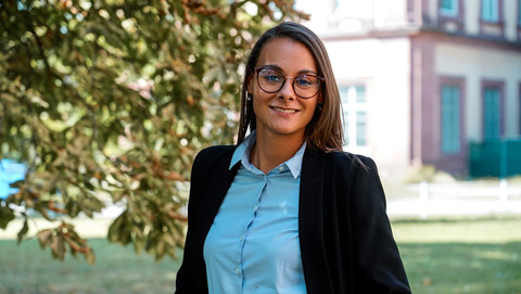 Yasmin Hoffmann. She is wearing a black jacket over a blue shirt and is standing on the Mensawiese with grass and trees in front of the castle.