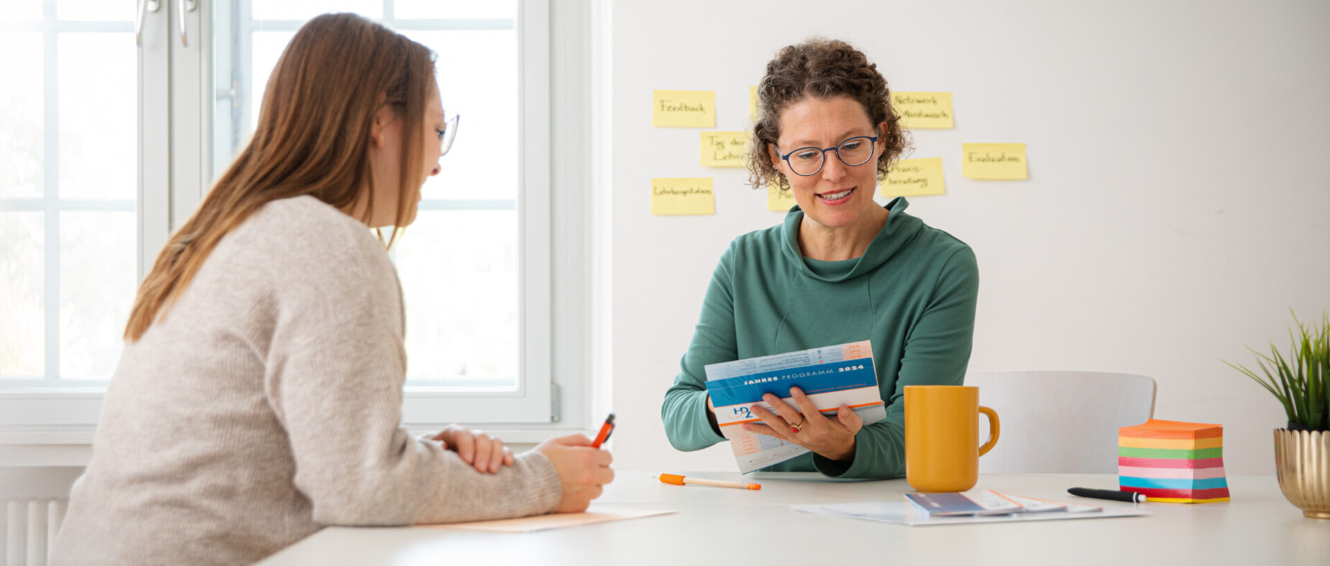 Zwei weiblich gelesene Personen sitzen an einem Tisch. Sie sind keine Studierenden. Eine Person hat eine Broschüre in der Hand. Beide betrachten diese und tauschen sich dazu aus.