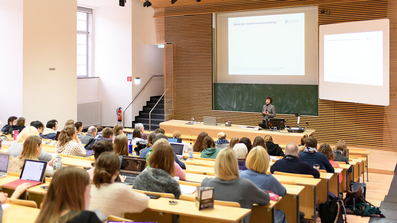 Studierende sitzen in einem großen Hörsaal. Vorne hält eine Dozentin einen Vortrag. Viele Studierende schreiben am Laptop.