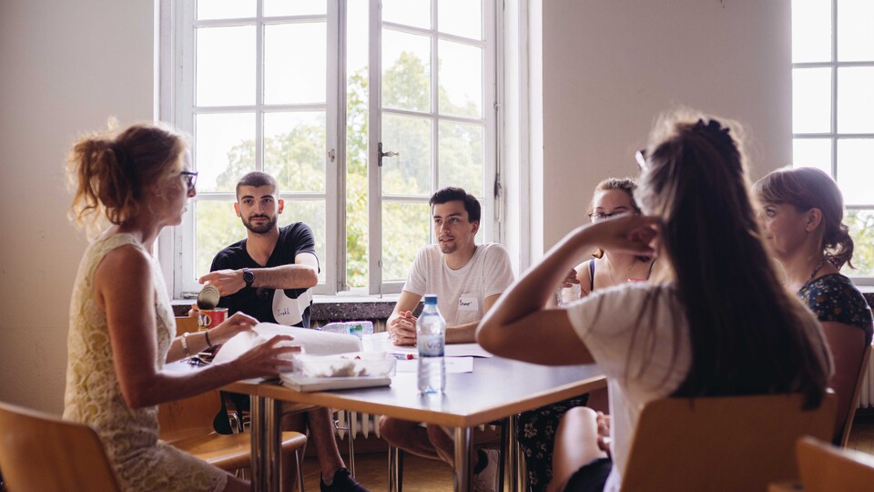 Sechs Personen sitzen um einen Tisch und führen ein Gespräch. Helle, freundliche Umgebung mit großen Fenstern.