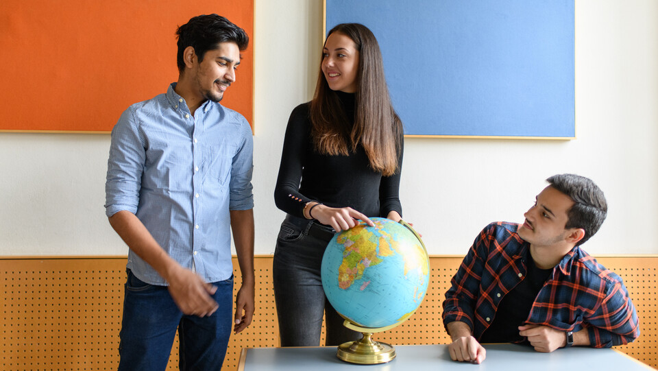 Drei Studierende an einem Tisch. Eine Studierende zeigt mit dem Finger auf einen Globus auf dem Tisch.