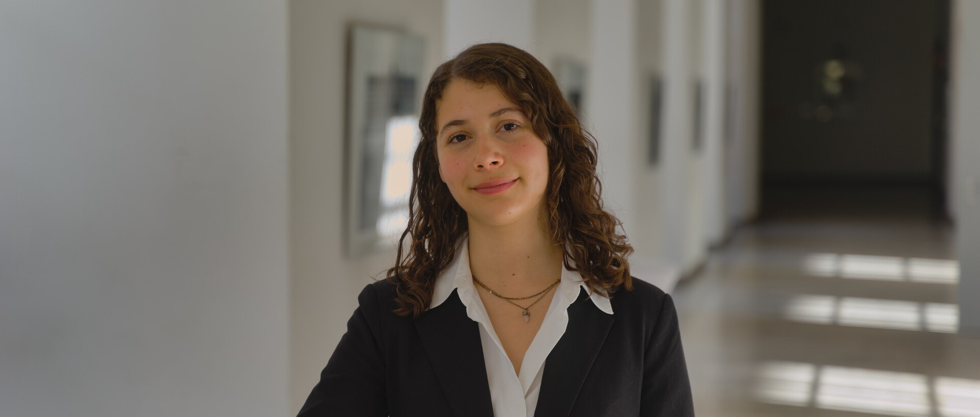Porträt von Emiliana. Sie steht in einem Gang der Universität, trägt eine weiße Bluse und ein schwarzes Jacket und schaut freundlich in die Kamera.