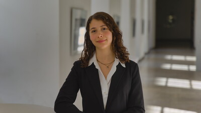 Porträt von Emiliana. Sie steht in einem Gang der Universität, trägt eine weiße Bluse und ein schwarzes Jacket und schaut freundlich in die Kamera.