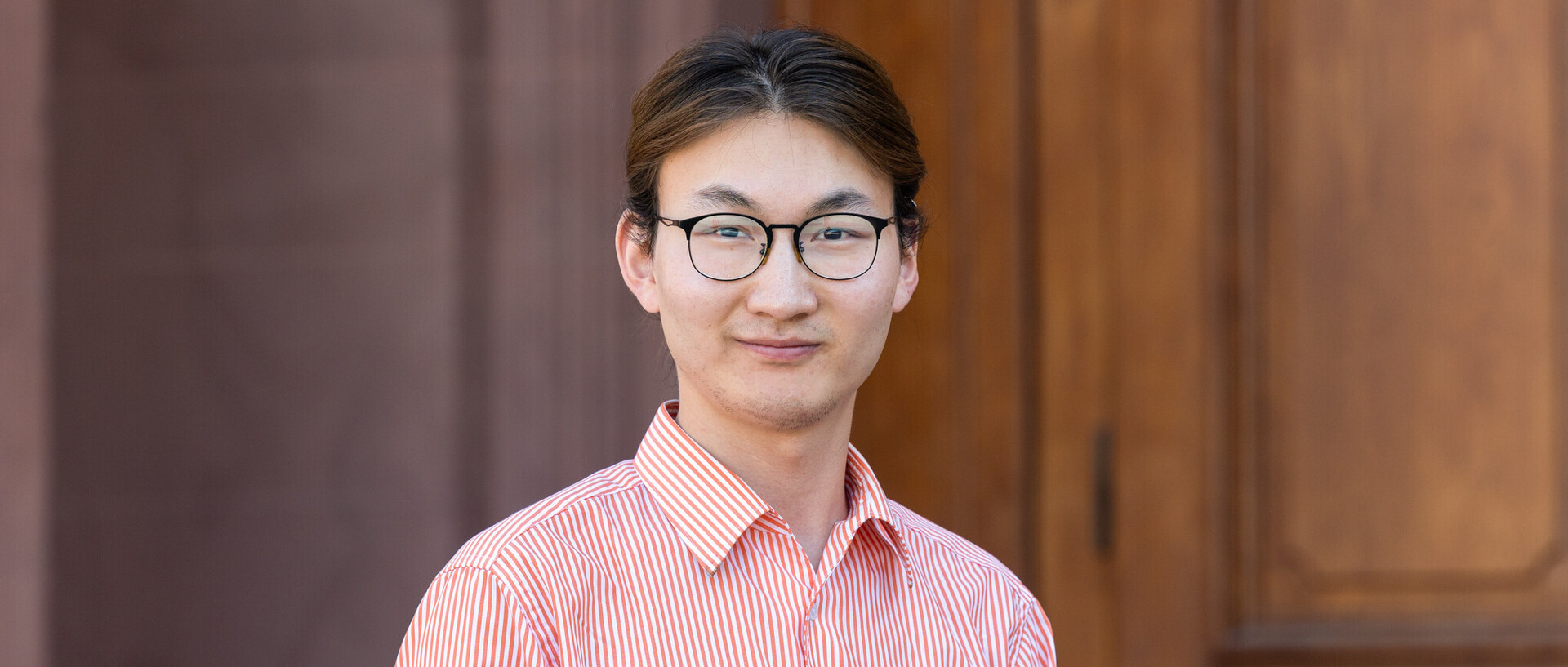 Khishigtugs hat lange braune Haare, die zu einem Zopf zusammengebunden sind und trägt eine Brille. Er hat ein rot-weiß gestreiftes Hemd an und lächelt in die Kamera.