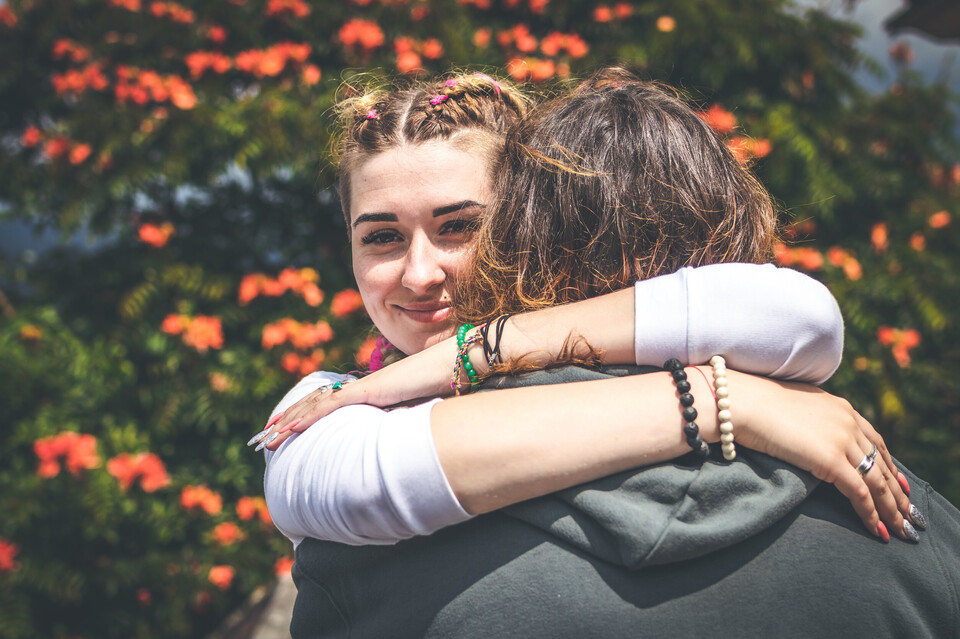 Zwei Personen umarmen sich. Eine Person steht mit dem Rücken zur Kamera, die andere Person lächelt.