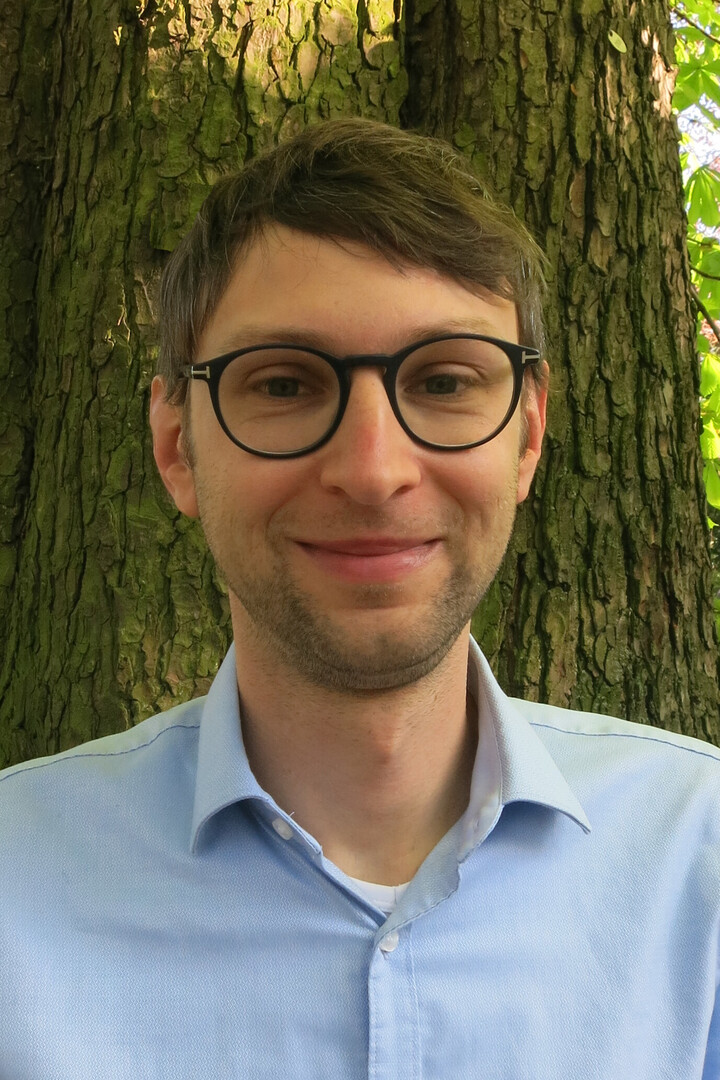 Eine lächelnde Person trägt ein hellblaues Hemd und steht vor einem Baum. Die Person heißt Roni Lehrer.