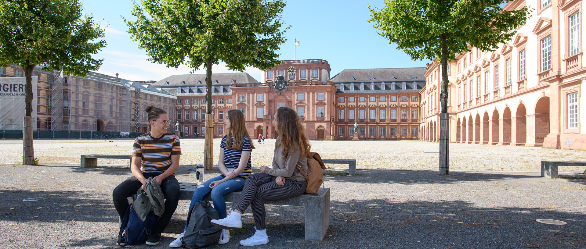 Drei studierende sitzen auf einer Steinbank im Ehrenhof. Hinter ihnen sieht man grüne Bäume und die Fassade des Schlosses.