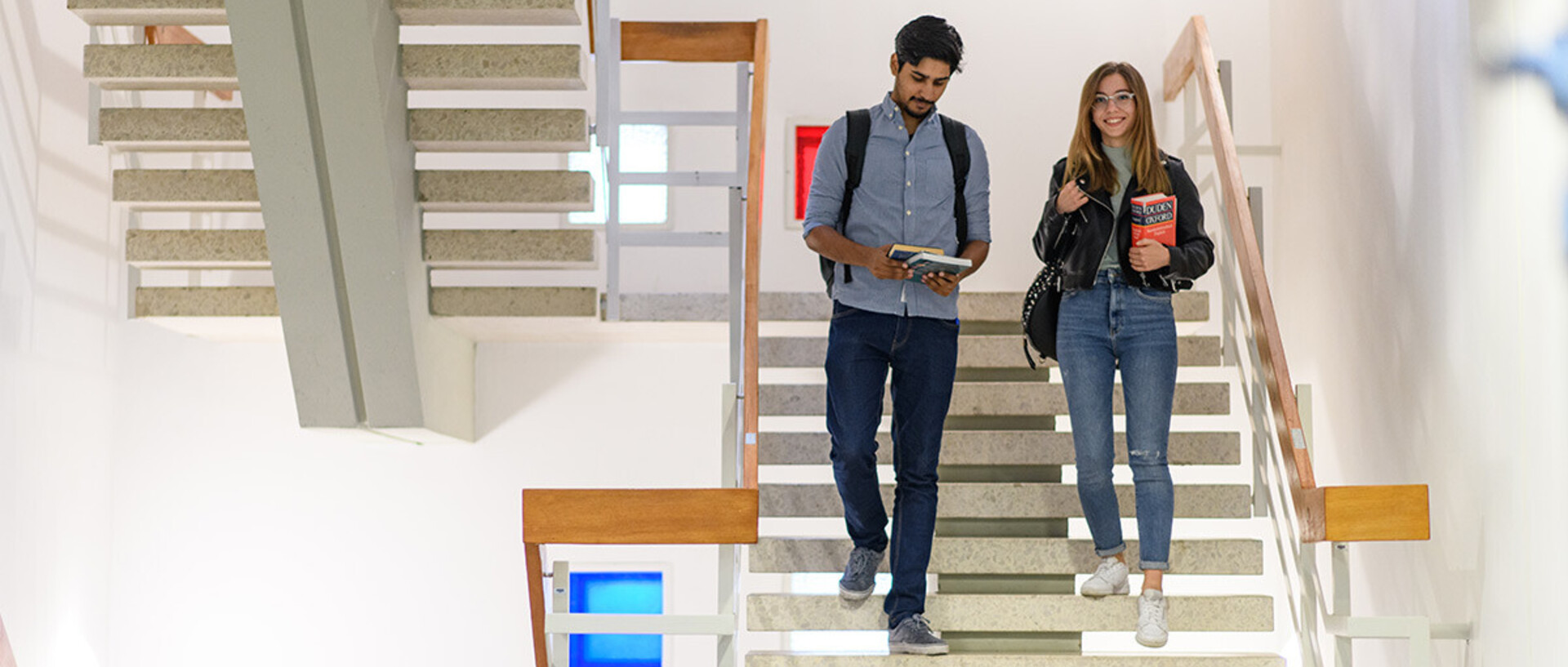 Eine Studentin und ein Student steigen eine helle Steintreppe herab. Sie halten Bücher in ihren Händen.