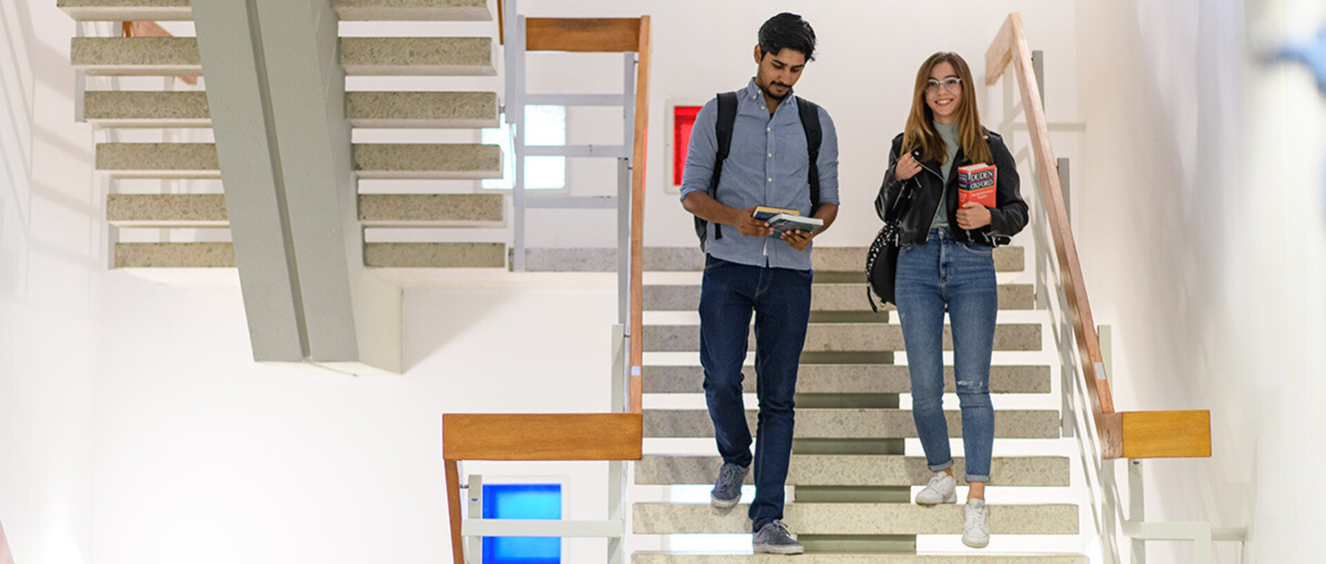 Eine Studentin und ein Student steigen eine helle Steintreppe herab. Sie halten Bücher in ihren Händen.