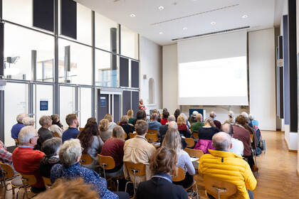 Viele Menschen sitzen in einem Saal auf stühlen und schauen auf eine Person, die spricht.