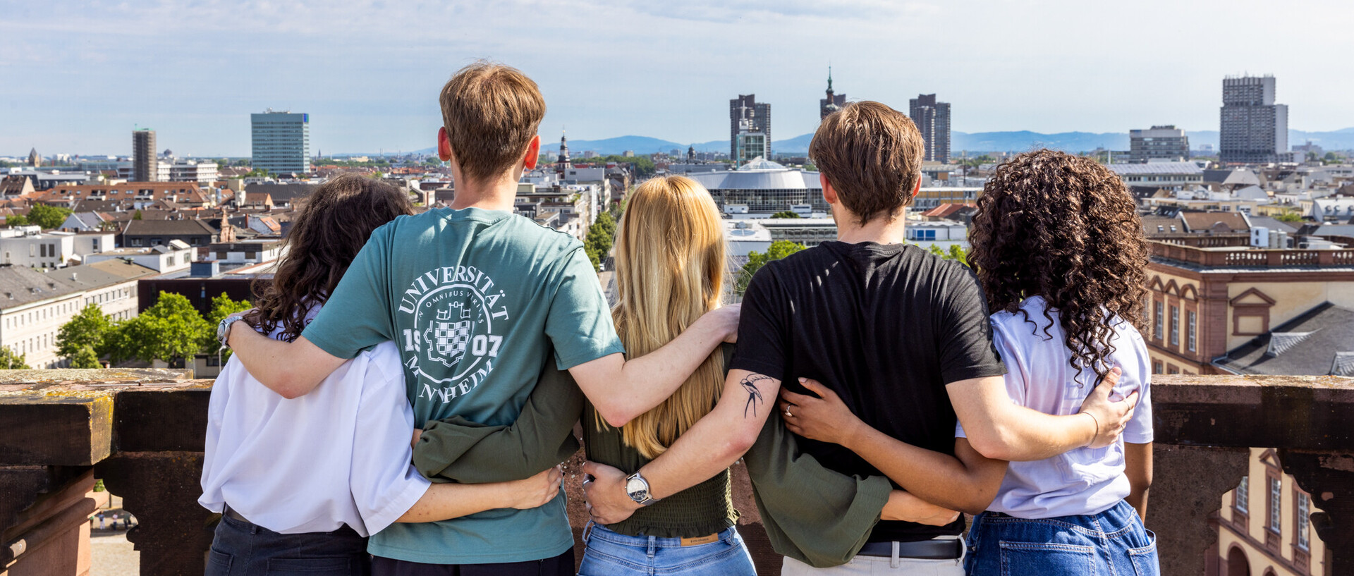 A group of five students on the roof of Schloss Mannheim. They all hold each other in their arms and take a look at the Ehrenhof or the city.