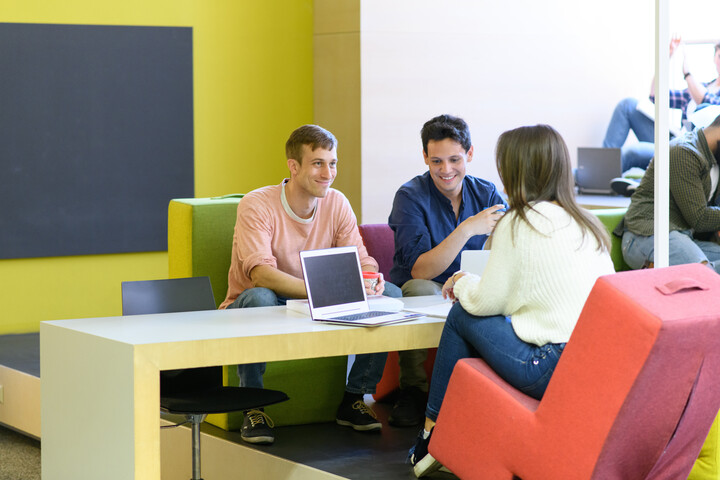 Drei Studierende sitzen gemeinsam an einem Tisch in einem modern gestalteten Lernraum und befinden sich im Austausch.
