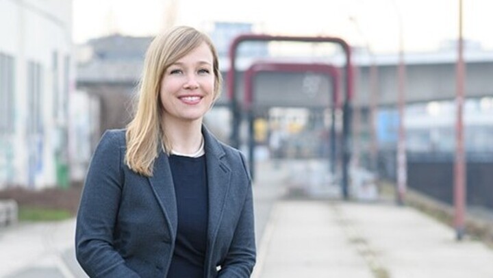Portrait von Julia Derkau. Sie trägt ein schwarzes Oberteil sowie einen grauen Blazer und steht vor einem Gebäude.