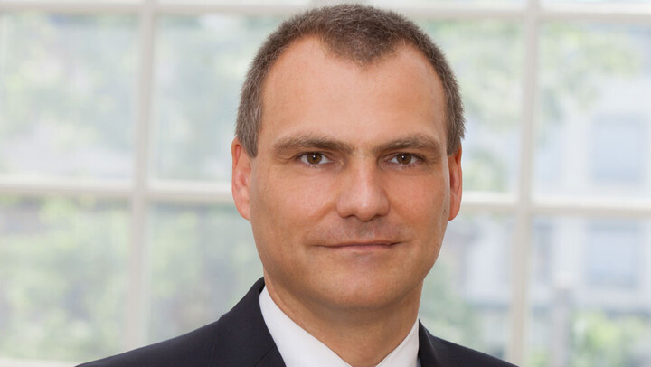 Portrait von Herr Simons. Er tägt einen Anzug und steht vor einer Fensterfront.