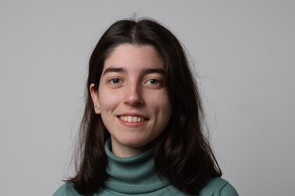 Rita Sousa hat dunkelbraune lange Haare und trägt einen hellgrünen Rollkragenpullover. Sie steht vor einem hellgrauen Hingtergrund.