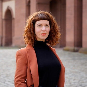 Corinna Kröger_Jula Jacob Jula Jacob hat lockige mittellange und rötliche Haare. Sie trägt einen kurze Pony und ein schwarzes Oberteil mit einem roten Blazer.