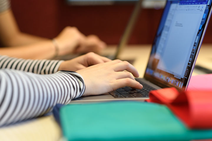 Eine Person, deren Arme und Hände im Bild sind, schreibt an einem Laptop. Sie trägt ein schwarz-weiß gestreiftes langärmeliges Oberteil. Im Hintergrund schreibt eine weitere Person an einem Laptop.