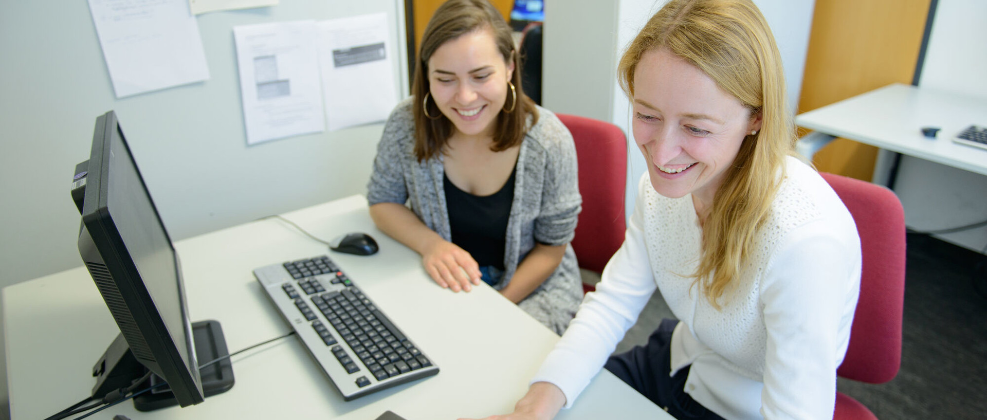 Zwei Studentinnen sitzen lachend an einem Tisch nebeneinander vor einem Laptop und einem Bildschirm mit Tastatur