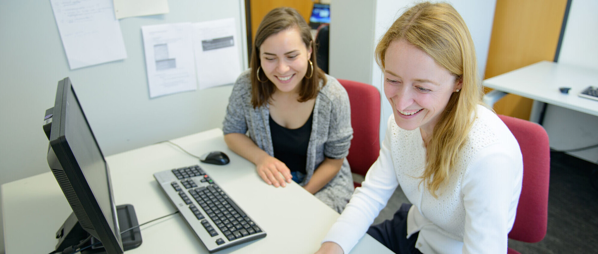 Zwei Studentinnen sitzen lachend an einem Tisch nebeneinander vor einem Laptop und einem Bildschirm mit einer externen Tastatur.