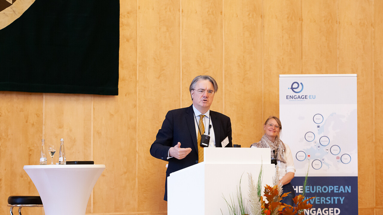 Eine Person trägt einen schwarzen Blazer und spricht hinter einem Rednerpult. Hinter ihm steht ein Banner von Engage.Eu.