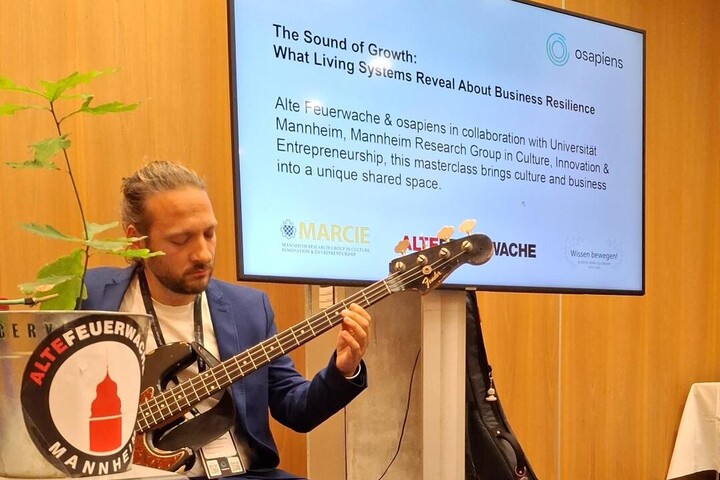 Mann mit Gitarre links im Bild, dahinter Powerpoint-Folie mit Titel: "The Sound of Growth: What Living Systems Reveal About Business Resilience". 