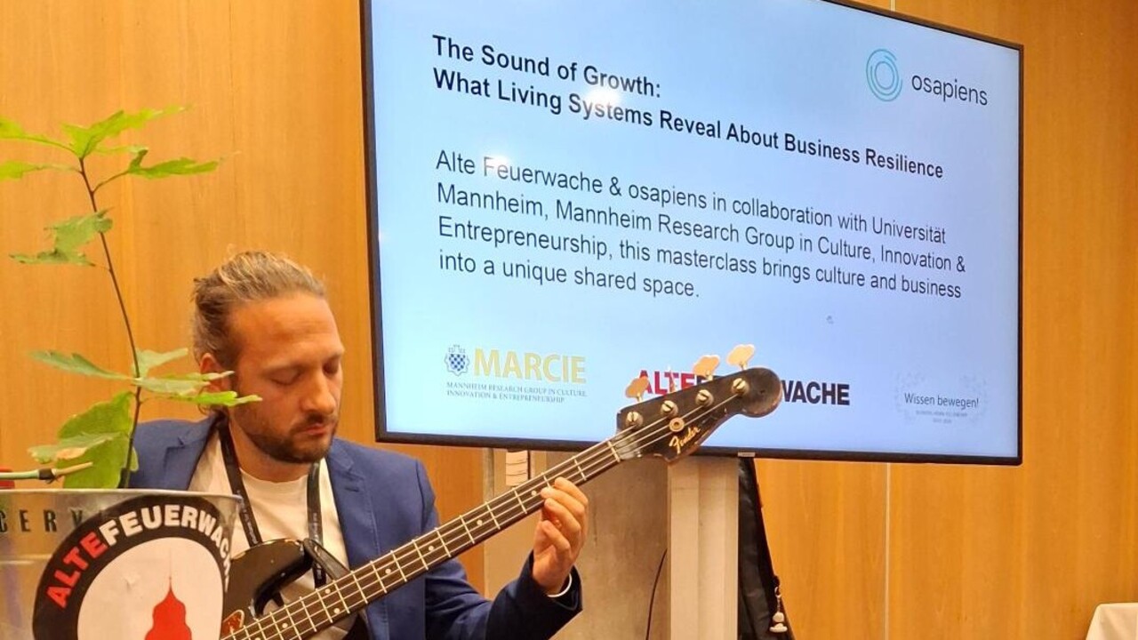 Mann mit Gitarre links im Bild, dahinter Powerpoint-Folie mit Titel: "The Sound of Growth: What Living Systems Reveal About Business Resilience".