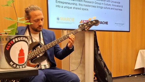 Mann mit Gitarre links im Bild, dahinter Powerpoint-Folie mit Titel: "The Sound of Growth: What Living Systems Reveal About Business Resilience".