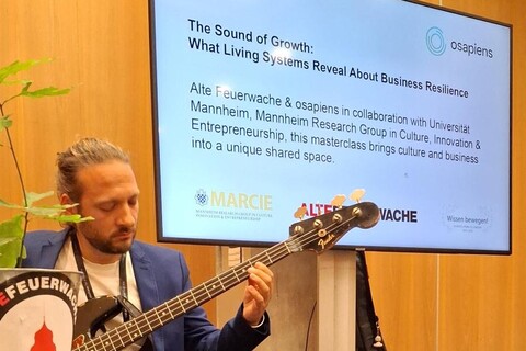 Mann mit Gitarre links im Bild, dahinter Powerpoint-Folie mit Titel: "The Sound of Growth: What Living Systems Reveal About Business Resilience".