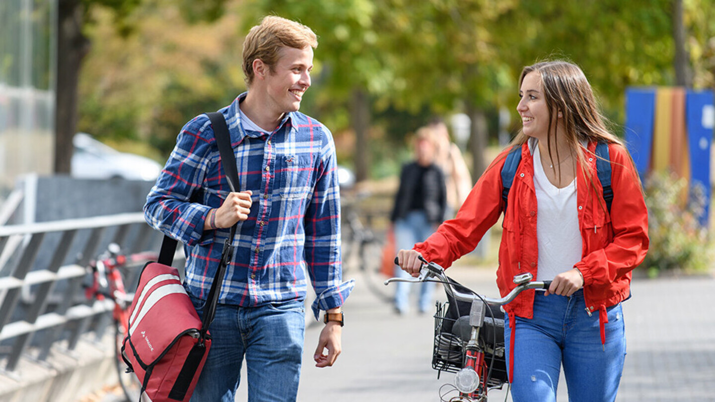 A female student and a male student laugh while walking side by side on a paved path. The female student is pushing her bike.