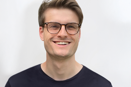 Porträt von Ulrich Roschitsch. Er trägt einen dunklen Pullover, hat dunkle Haare und eine Brille. Er lacht in die Kamera.