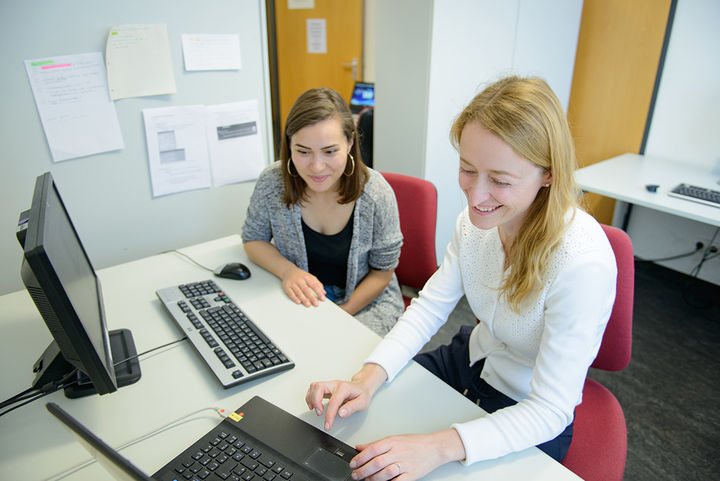 Zwei Personen an Büroarbeitsplätzen mit Computern im Gespräch.