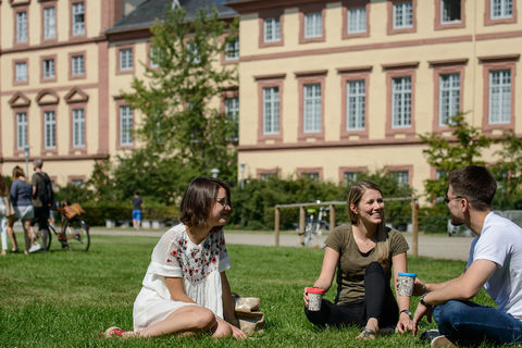 Zwei Studentinnen und ein Student sitzen auf einer grünen Wiese. Sie tragen frühlingshafte Kleidung und halten Kaffee-To-Go-Becher in ihren Händen.