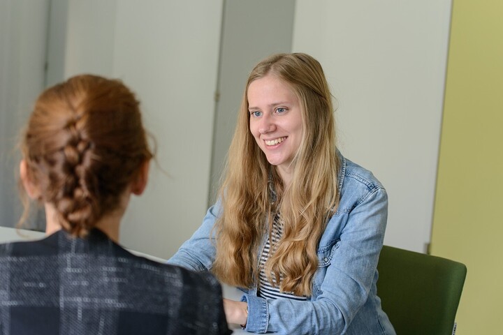 Zwei Personen sitzen sich an einem Tisch gegenüber. Eine junge Frau lächelt und wirkt im Gespräch.