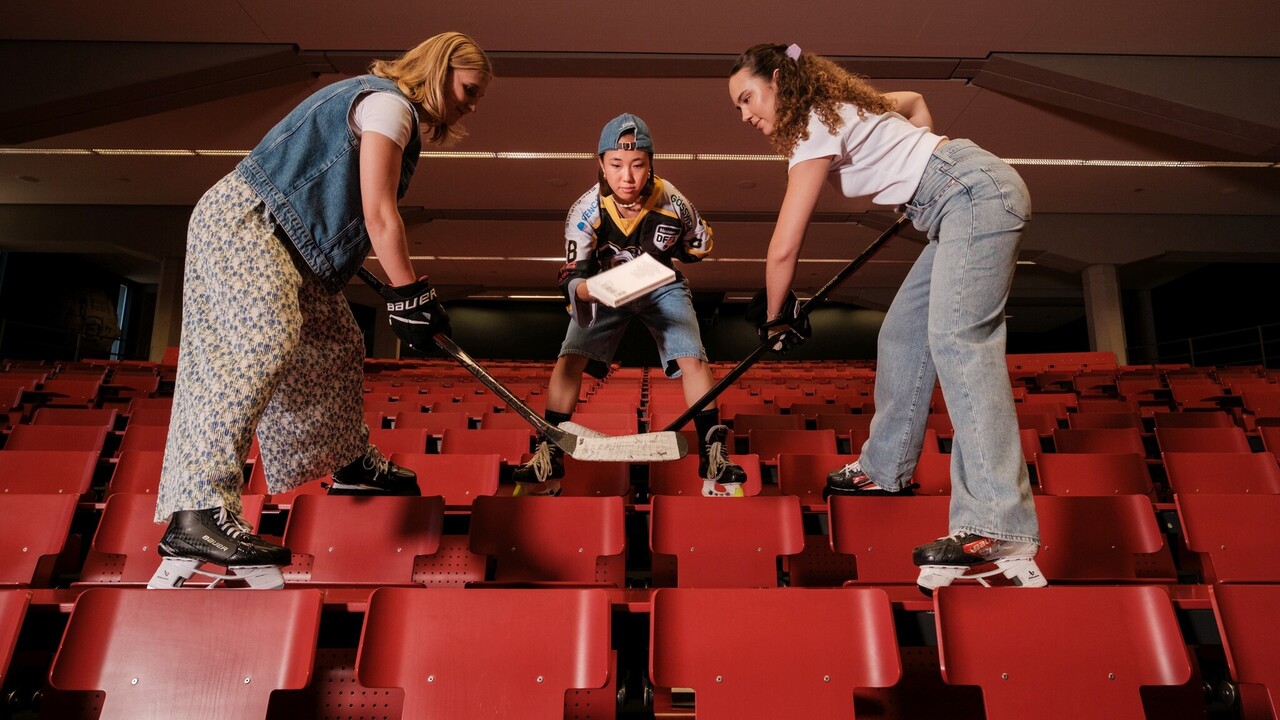 Auf den Tischen eines Vorlesungssaals stehen drei Eishockeyspielerinnen. Zwei halten ihre Schläger in Startposition, die mittlere hält ein Buch.