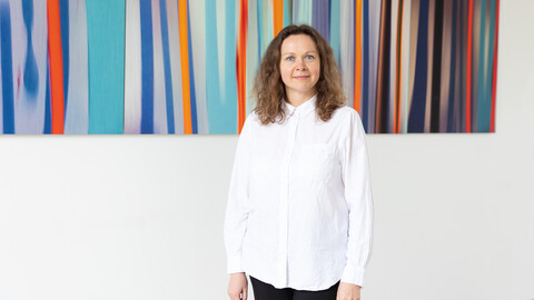 Eine Person trägt ein weißes Hemd sowie eine schwarze Hose und steht vor einem bunten Gemälde. Die Person heißt Melina Liulikova.