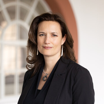 Eine Person trägt eine schwarze Bluse sowie einen schwarzen Blazer und steht vor einem Fenster. Die Person heißt Kirstin Kersten.