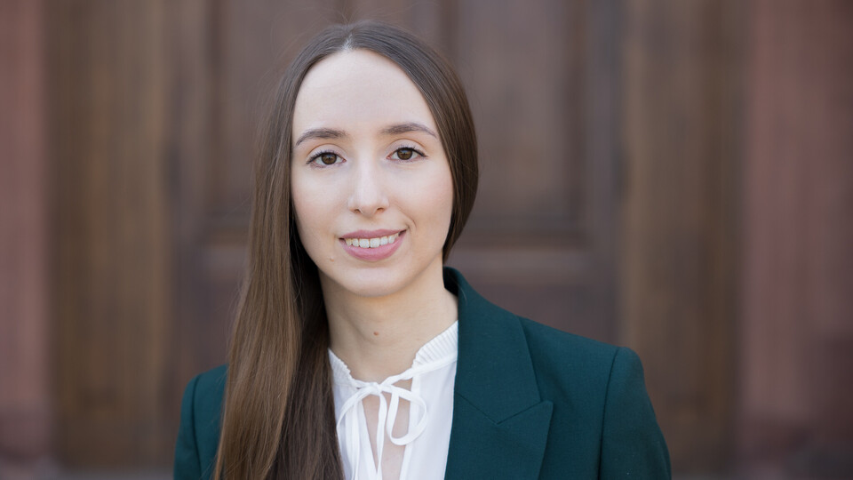 Sabine Stillger. Sie trägt ein graues Jackett über einem weißen Oberteil und steht vor einer Holztür.