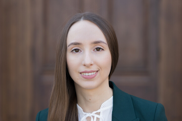 Sabine Stillger. Sie trägt ein graues Jackett über einem weißen Oberteil und steht vor einer Holztür.