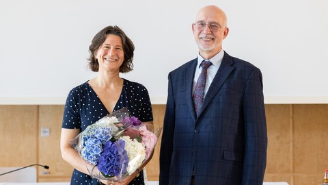 Zwei Personen in schicker Kleidung stehen nebeneinander. Die Person links, Katrin Schoppa-Bauer, hält einen Blumenstrauße in ihren Händen.