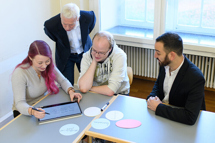 Eine junge Frau mit pinken Haaren zeigt drei interessierten Männern eine Skizze auf ihrem Laptop.