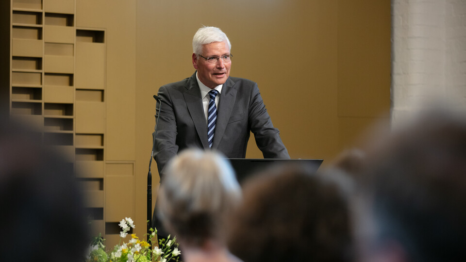 Eine Person mit kurzen grauen Haaren, einer Brille und einem grauen Anzug steht an einem Rednerpult cor Menschen, die verschwommen von hinten zu sehen sind