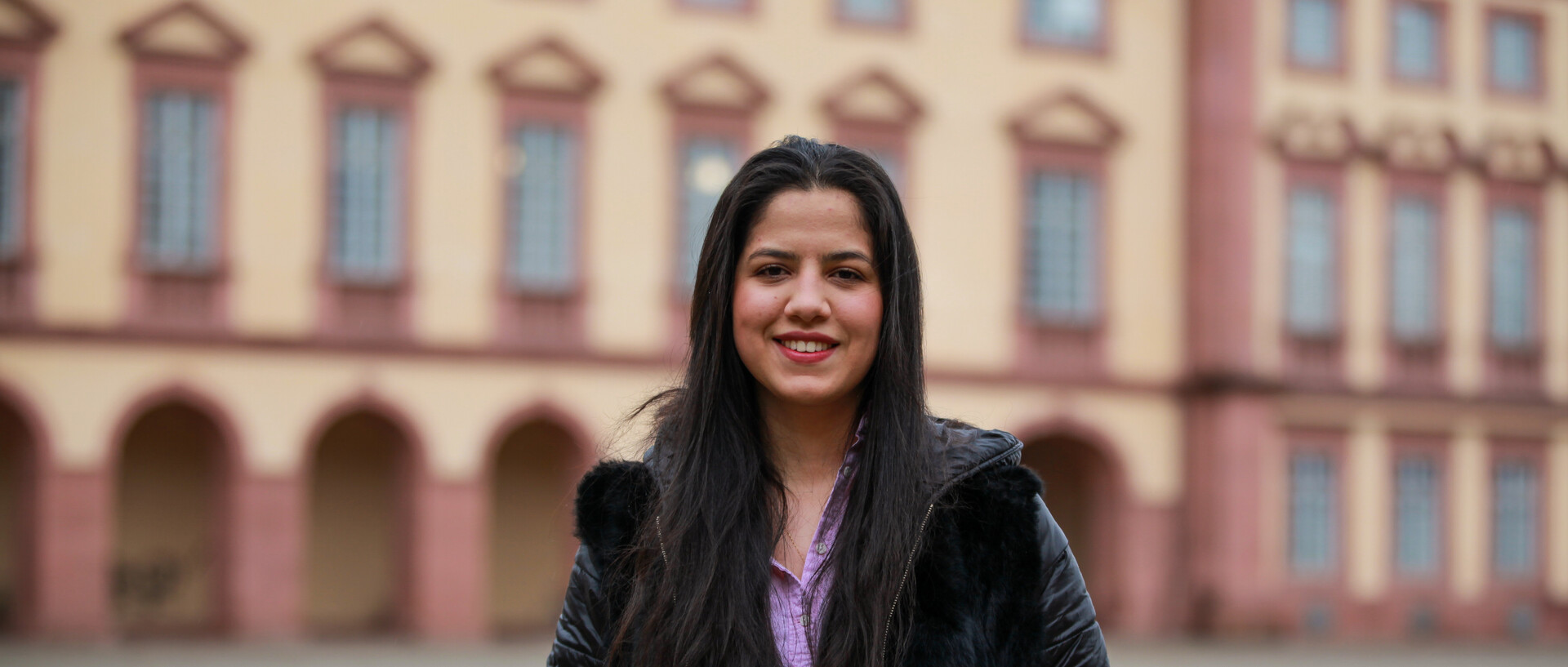 Eine lächelnde Person trägt eine lila-farbene Bluse sowie eine schwarze Jacke und steht vor dem Schloss. Die Person heißt Hala Fayad.