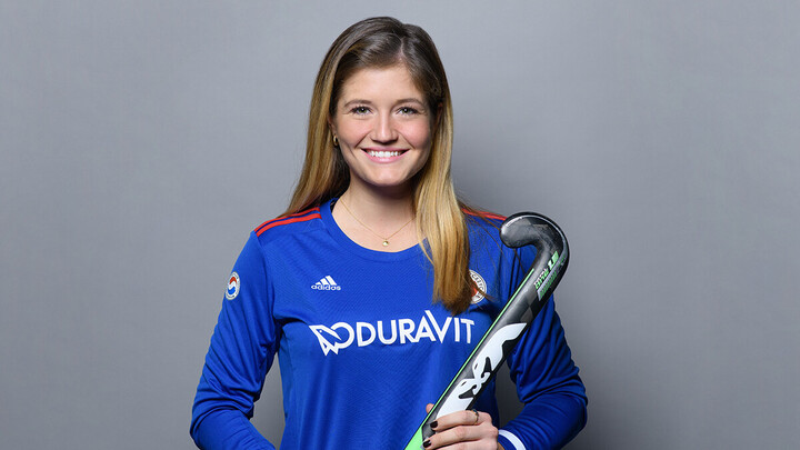 Sonja Zimmermann steht lächelnd vor einer grauen Wand. Sie trägt ein blaues Trikot und hält vor sich einen Hockeyschläger.