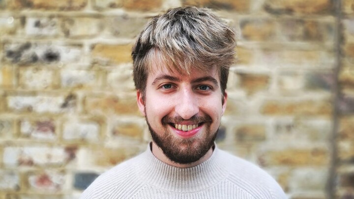 Johannes Lattmann. He is wearing a white sweater and is standing in front of brownish brickwall.