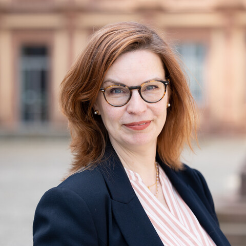 Sabine Gehrlein im Ehrenhof des Mannheimer Schlosses. Sie trägt eine Brille, eine gestreifte Bluse und einen dunklen Blazer darüber.