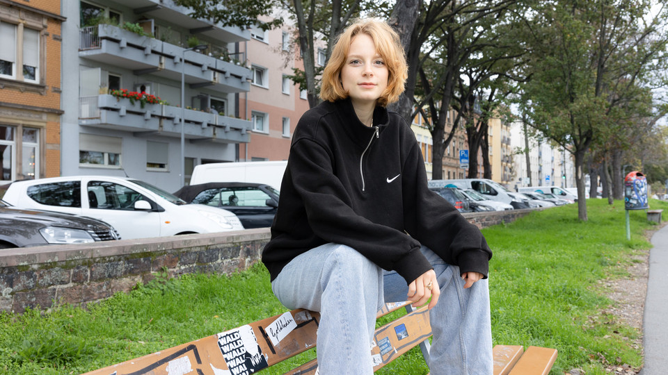 Eine Studentin mit kurzen rot-blonden Haaren sitzt auf der Lehne einer Bank in der Neckarstadt-West.