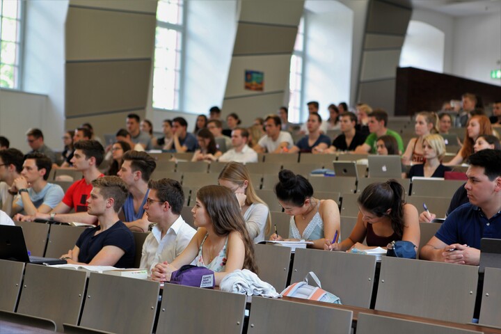 Studierende sitzen in einem Hörsaal. Die Studierenden tragen kurzärmlige Oberteile und vor ihnen liegen Laptops, Tablets oder Notizblöcke.
