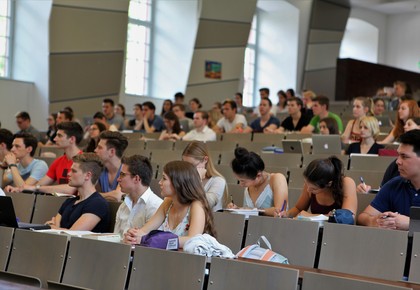 Studierende sitzen in einem Hörsaal. Die Studierenden tragen kurzärmlige Oberteile und vor ihnen liegen Laptops, Tablets oder Notizblöcke.